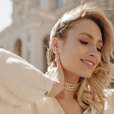 Cheerful blonde curly woman in pearl necklace and white elegant blouse smiles sincerely, looks down and poses outside.