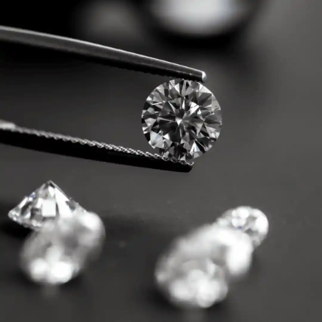 Tweezers holding a brilliant round diamond above four other loose gemstones.